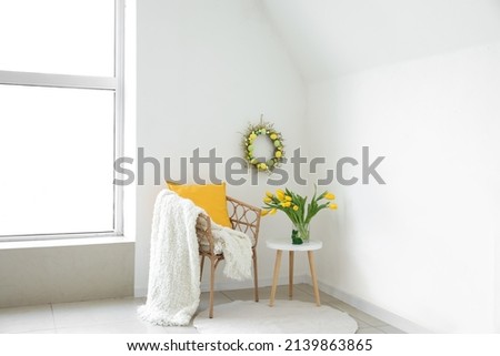 Chair and table with bouquet of tulips near light wall with handmade Easter wreath in room