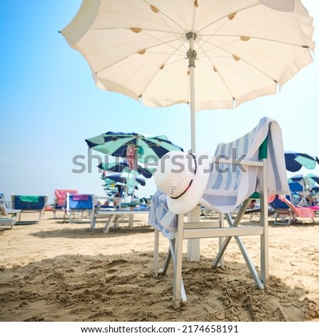 Chair with white straw hat and striped towel under a beach umbrella on a sunny summer day. In the background, the beach equipped with sunbeds and umbrellas along the Adriatic Sea in Italy.