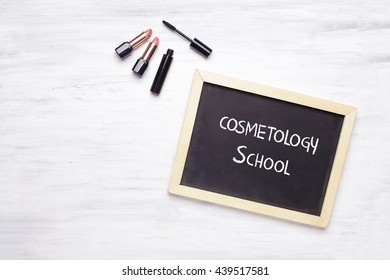 Chalkboard With Cosmetology School Written On It, And Cosmetic Products On White Wooden Background. Fashion And Makeup Concept.