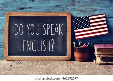 a chalkboard with the question do you speak English? written in it, a pot with pencils and the flag of the United States, on a wooden desk