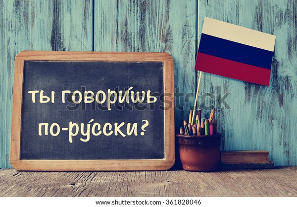 a chalkboard with the question do you speak\
russian? written in russian, a pot with pencils, some books and the\
flag of Russia, on a wooden\
desk
