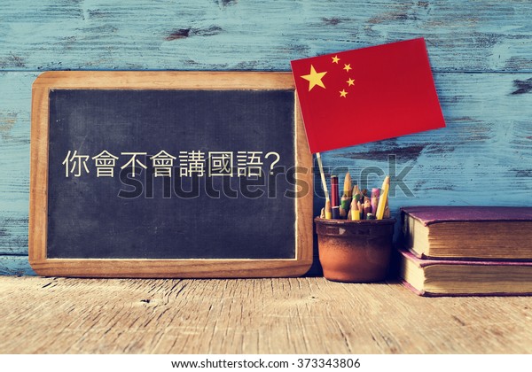 a chalkboard with the question do you speak\
chinese? written in chinese, a pot with pencils, some books and the\
flag of china, on a wooden\
desk