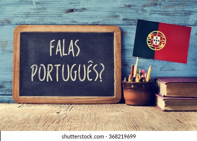 a chalkboard with the question question falas portuges? do you speak Portuguese? written in Portugese, a pot with pencils, some books and the flag of Portugal, on a wooden desk