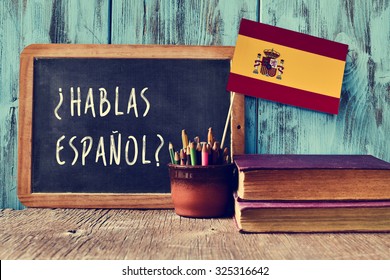 a chalkboard with the question hablas espanol? do you speak Spanish? written in Spanish, a pot with pencils and the flag of Spain, on a wooden desk