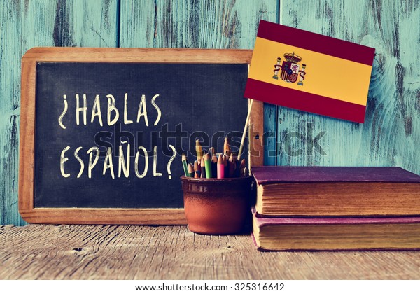 a chalkboard with the question hablas\
espanol? do you speak Spanish? written in Spanish, a pot with\
pencils and the flag of Spain, on a wooden\
desk