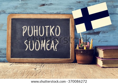 a chalkboard with the question puhutko suomea, do you speak Finnish? written in Finnish, a pot with pencils, some books and the flag of Finland, on a wooden desk