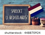 a chalkboard with the question Spreekt u Nederlands?, do you speak Dutch? written in Dutch, a pot with pencils, some books and the flag of the Netherlands on a wooden desk