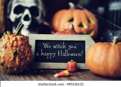 a chalkboard with the text We witch you a happy Halloween, surrounded by some different pumpkins, placed on a rustic wooden surface, and some scary ornaments, such as a skull or a carved pumpkin