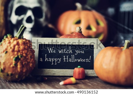 a chalkboard with the text We witch you a happy Halloween, surrounded by some different pumpkins, placed on a rustic wooden surface, and some scary ornaments, such as a skull or a carved pumpkin