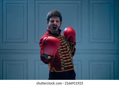 Challenge to a duel. Emotional young man like medieval hussar wearing boxing gloves isolated on dark blue background. Sport, retro style, hobby, comparison of eras concept.