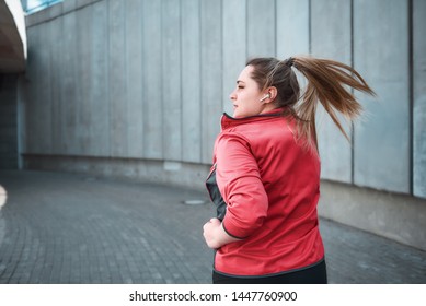 Challenging myself. Back view of young attractive plus size woman in sporty clothes is looking aside and listening music while running in the city - Powered by Shutterstock