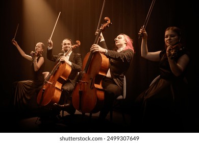 Chamber quartet of string musicians sitting in row on stage with project light raising bows while thanking public at end of performance - Powered by Shutterstock