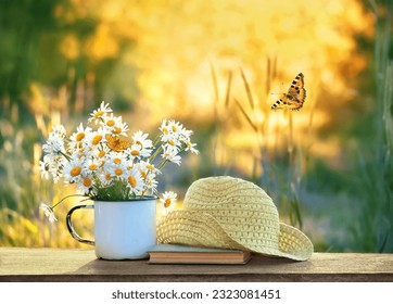 chamomile flowers in cup, butterflies, book and braided hat on table in garden, sunny natural abstract background. summer season. Beautiful rustic floral composition. relaxation, harmony atmosphere.