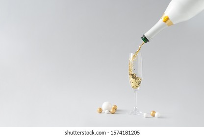 Champagne bottle pouring golden glitter into the glass with white and golden decoration against grey background. Celebration minimal Christmas party.