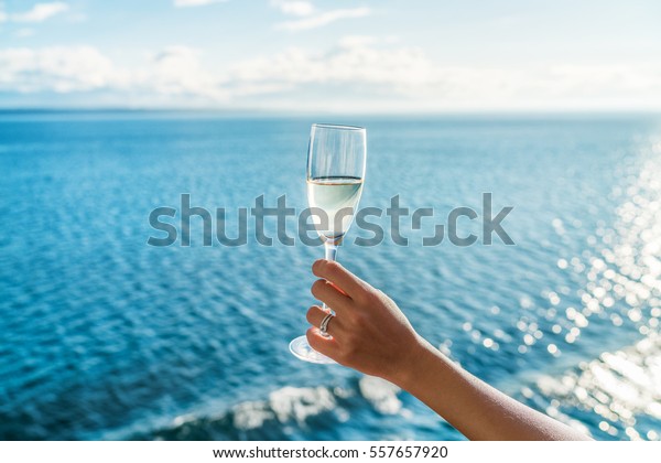 Champagne glass woman\'s hand\
toasting on ocean background at luxury cruise ship during sunset.\
Travel vacation for honeymoon, lady holding flute wearing wedding\
ring.