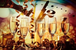 Champagne Glasses And Clock At Twelve Against Holiday Lights - New Year's Eve
