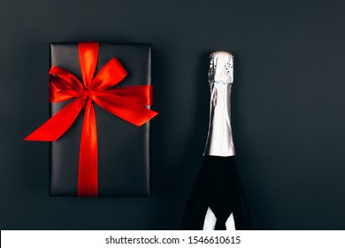 Champagne With Present Box On Dark Background. Festive Concept. Flat Lay Style. Black Box With Red Bow.