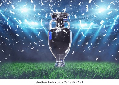The champion cup stands on a green field at a football stadium with the light of spotlights and confetti. Football cup final, creative idea. Euro 2024 celebration