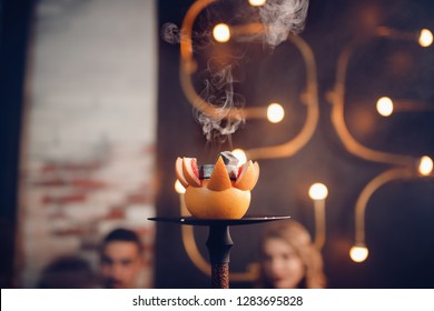 Change Coals And Tobacco Hookah On Fruit. In Background Group Of Friends People Are Resting And Smoking Shisha.