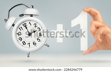 Change time. Spring forward, on a gray background. A white alarm clock indicates that the time has been moved forward an hour. Hand holding number one with plus sign, Spring forward concept
