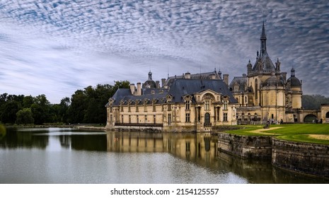 Chantilly, France, 08,26,2021: Panoramic View Of The Château De Chantilly Reflected In The Water Of The River With Light Clouds In The Sky.