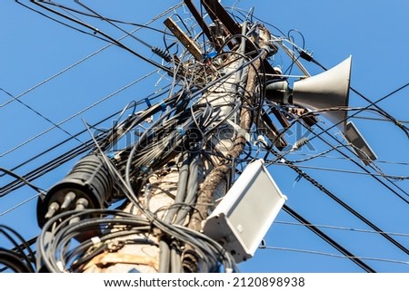 Chaos of many different data cables and network wires messy tangled hanged on concrete pillar pole on urban city street against clear blue sky on day time. Announcement speaker mounted on column