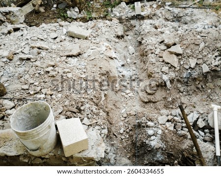 A chaotic construction site featuring a white plastic bucket with cement residue beside a rough concrete block. The ground is strewn with dirt, rocks, and debris, showcasing a raw, industrial scene.