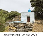 Chapel, small whitewashed stonewall Greek Orthodox Church with cross and wooden door on rocky landscape. Tinos island religious destination.