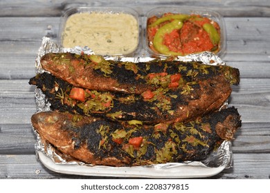 Charcoal Grilled Mullet Fish With Parsley And Tomato, Pickled Eggplant Aubergine And Tahini Sauce, Barbecued And Served In A Disposable Plate On Foil Paper Isolated On Wooden Background