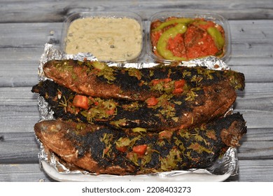 Charcoal Grilled Mullet Fish With Parsley And Tomato, Pickled Eggplant Aubergine And Tahini Sauce, Barbecued And Served In A Disposable Plate On Foil Paper Isolated On Wooden Background