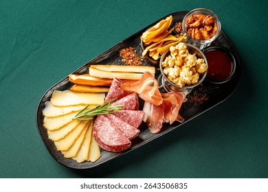 Charcuterie board with cheese, cured meats, crackers, popcorn, nuts, and dipping sauce, artistically arranged on a black platter over a green surface. Gourmet snack concept.