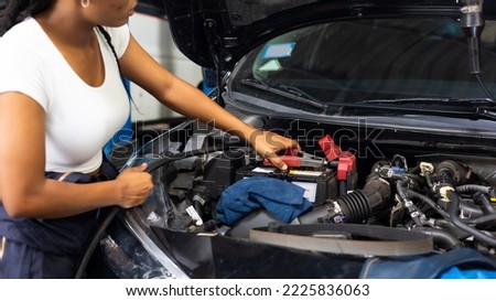 Charge electric power to ca battery by charging jumper cables. African  mechanic female uses  multimeter voltmeter to check voltage level in car battery at car service and maintenance garage.