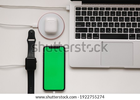 Charging of different devices on working place. Few cabel for wireless charging. Laptop, smart watch, smart phone with green screen and witeless headphones on white table. Top view. Mock up. Place for
