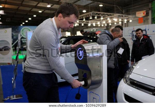 Charging electric car:\
male driver holding electric charger pluged in a charging station.\
Exhibition PLUG-IN UKRAINE 2018. March 2, 2018. Kiev Expo center.\
Kiev, Ukraine\
