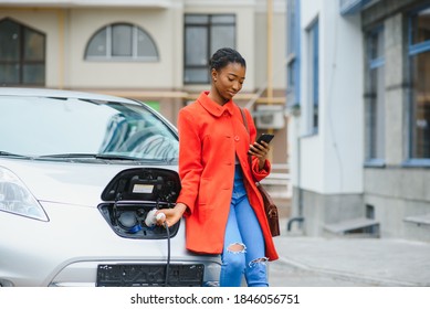 Charging Electro Car At The Electric Gas Station. African American Girl Standing By The Car.