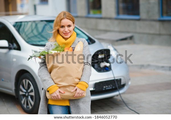 Charging electro car at the electric\
gas station. Woman standing by the car. Lady with\
foodstuff.