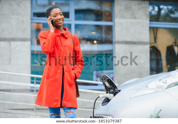 Charging electro car at the electric\
gas station. African American girl standing by the\
car.