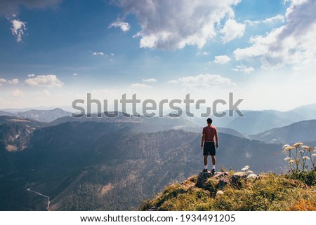 Charging energy in wild Austrian nature. Athlete of lighter figure with cap and red elastic trick stands on the edge of rock and enjoys feeling of relaxation and freedom. Conquering Mount Otscher.