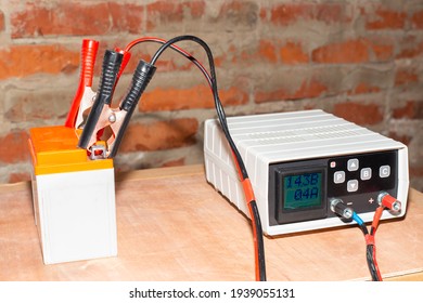Charging The Motorcycle Battery. On A Wooden Surface Against The Background Of A Brick Wall Of The Garage, The Battery Of A Motorbike Is Charged With A Charger.