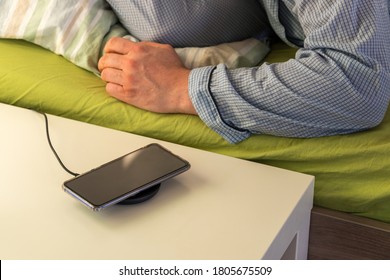 A Charging Phone On The Bedside Table Next To The Bed And A Sleeping Man