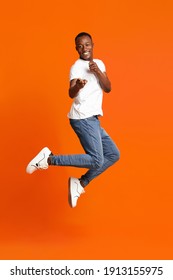 Charismatic Black Guy Pointing At Camera While Jumping Up On Orange Studio Background. Happy African American Young Man Indicating At Camera, Cheerfully Flirting And Bouncing