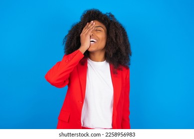 Charismatic carefree joyful young businesswoman with afro hairstyle wearing red over blue wall likes laugh out loud not hiding emotions giggling hear funny hilarious joke chuckling facepalm.