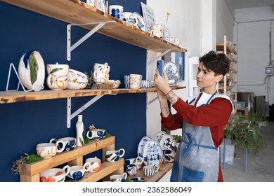 Charismatic Caucasian woman seller in souvenir shop takes pictures of goods at phone for posting on social networks. Successful girl store employee with ceramic tableware shoots video on smartphone - Powered by Shutterstock