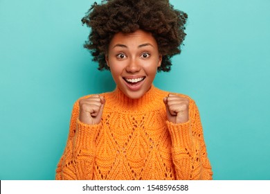 Charismatic cute African American lady cheers victory, raises clenched fists, celebrates something, smiles broadly, shows white teeth, wears knitted orange sweater, being extremly excited feels upbeat - Shutterstock ID 1548596588