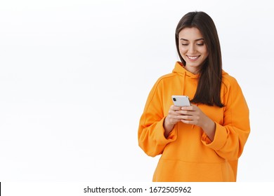 Charismatic Pretty Brunette Girl In Orange Hoodie Smiling And Looking Mobile Phone Screen As Texting, Messaging With Boyfriend, Using Phone Application, Edit Pics With Funny Filters, White Background