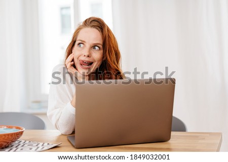 Charismatic young woman with a look of anticipation glancing eagerly to the side with her tongue out as she sits at a laptop computer at a table