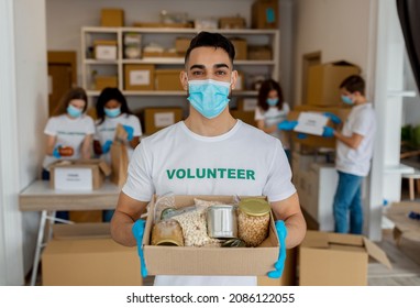 Charity Center. Arab Activist Male Volunteer Holding Food Donation Box And Looking At Camera, Wearing Medical Face Mask. Man Working At Charity Organization Center With Friends