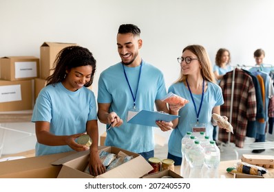 Charity Center Work. Young Male Manager Checking Donation List And Writing In Clipboard, Multiracial Volunteers Working In Charity Organization, Packing Donations Boxes