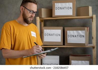 Charity Center Work. Young Male Manager Checking Donation List And Writing In Clipboard, Working In Charity Organization, Packing Donations Boxes. Copy Space