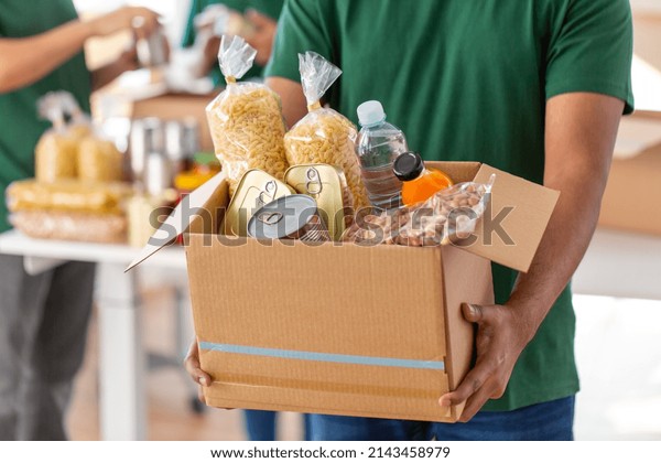 charity, donation and\
volunteering concept - close up of male volunteer\'s hands holding\
box with food over group of people at distribution or refugee\
assistance center
